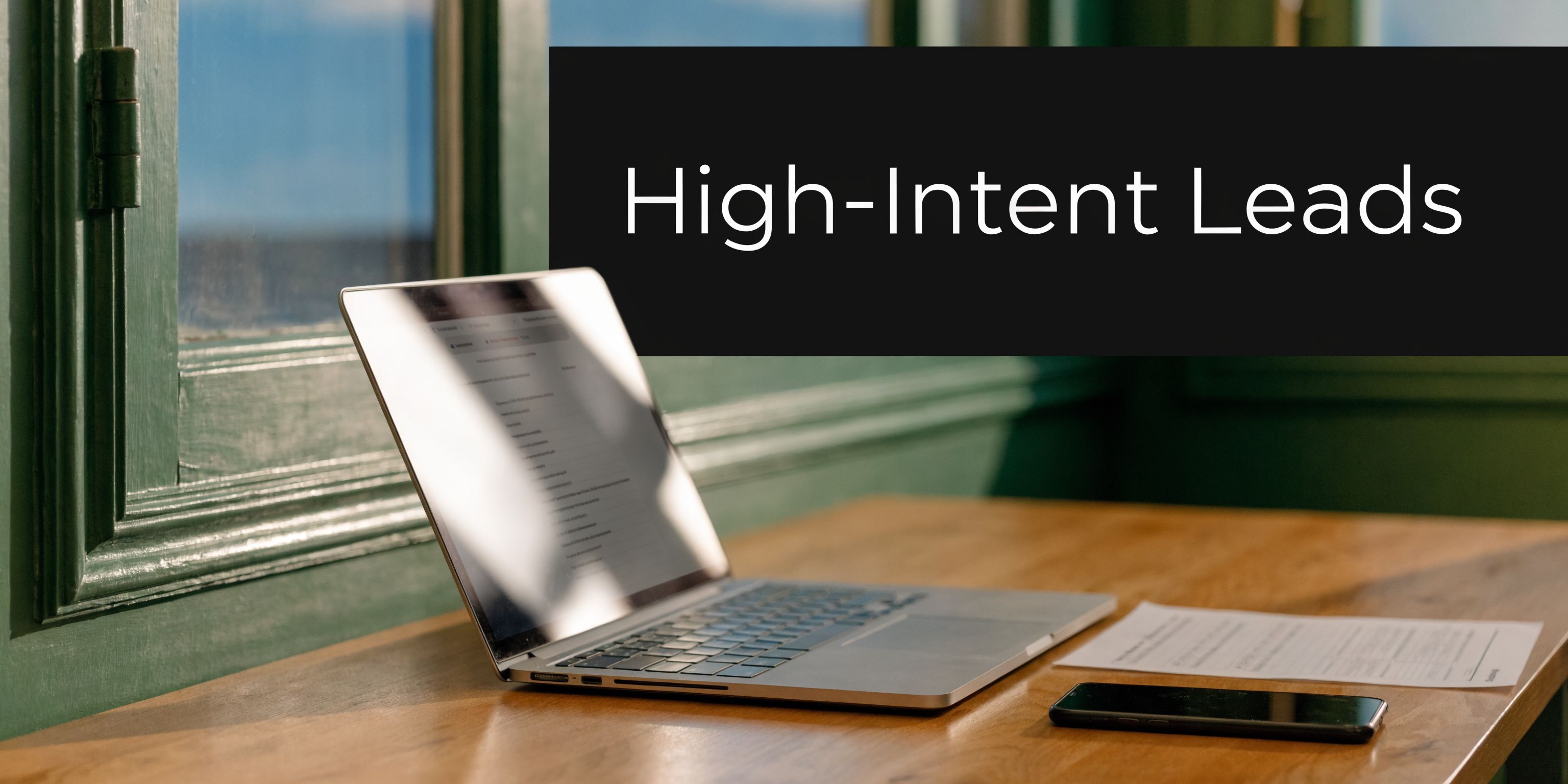 A modern laptop on a wooden desk next to a smartphone and paper, with high-intent leads text.