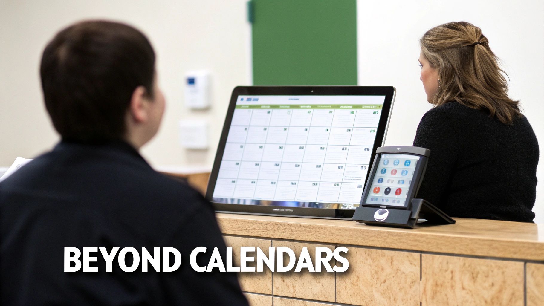 Two people in an office setting looking at a large digital calendar display and a smaller device.