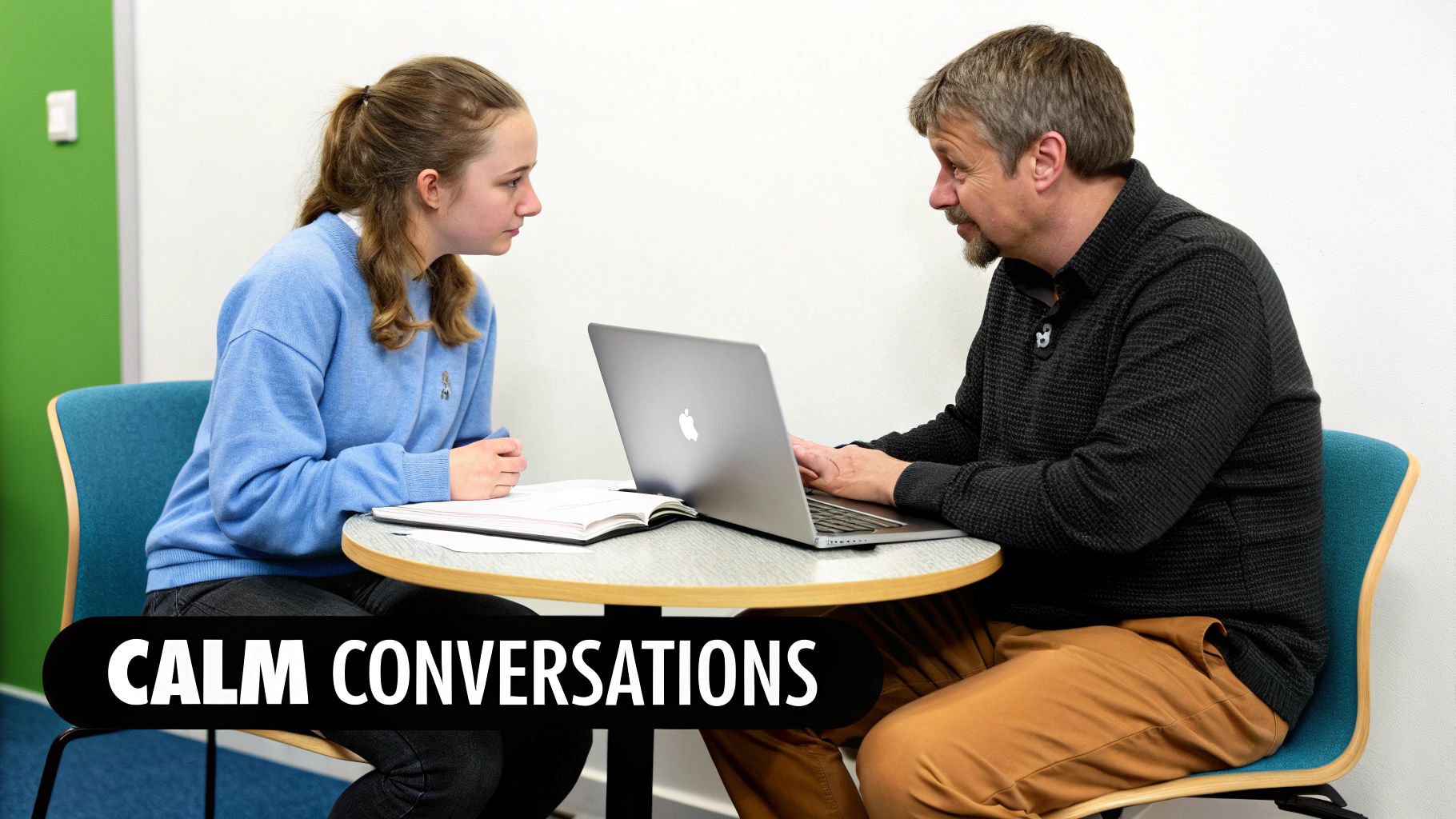 A man and a young woman sit at a table with a laptop, engaged in a calm conversation.