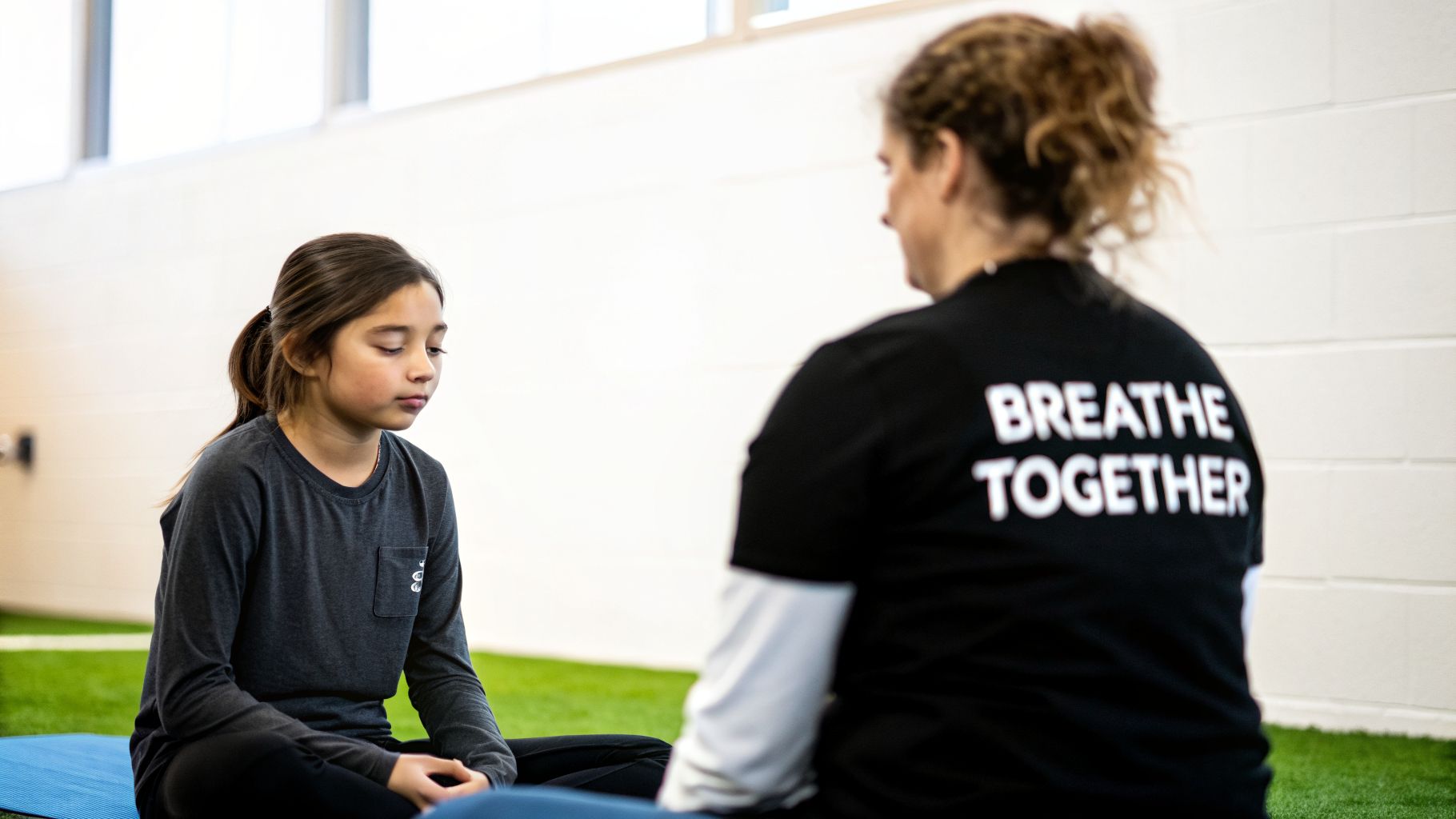 A young girl meditates on a blue mat, facing an adult wearing a "BREATHE TOGETHER" shirt.