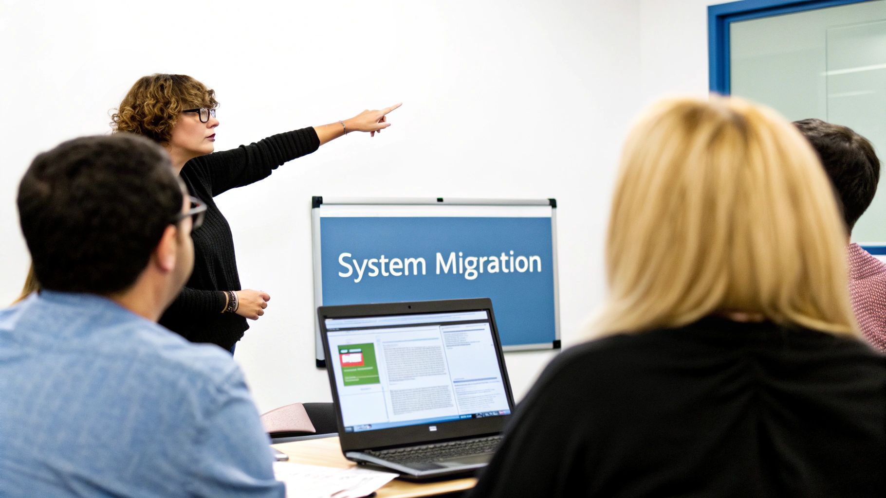 A woman with curly hair and glasses presents on 'System Migration' to an audience in a meeting.